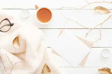 Örgü ekose ile rahat ev masası masa, çay bardağı, sonbahar yaprakları, ahşap beyaz arka plan üzerinde boş kağıt kartı ile mektup. Üst görünüm, düz döşeme, kopyalama alanı. Sonbahar kompozisyonu. İskandinav hygge tarzı kavramı.