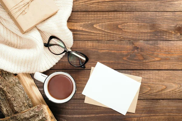 Beyaz örme eşarp, çay bardağı, yakacak odun, mektup, boş kağıt kart, ahşap masa üzerinde gözlük ile Flatlay kompozisyon. Hygge tarzı, rahat sonbahar veya kış konsepti. Düz döşeme, üst görünüm, genel merkez.