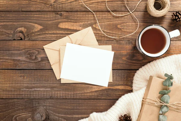 Sonbahar flatlay kompozisyon. Çerçeve ahşap arka plan üzerinde zanaat kağıt zarf, boş kart, çay bardağı, ip, hediye kutusu, beyaz örme eşarp yapılmış. Sonbahar hygge tarzı masa veya kış tatili konsepti.