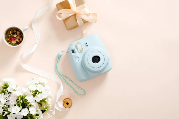 Modern instant camera, daisy flowers, cup of tea, gift box, ribbon on pastel pink background. Flat lay, top view, overhead. Fashion blogger workspace, beauty and technology concept.