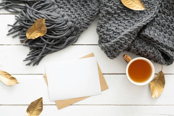 Vista dall'alto accogliente scrivania con plaid lavorato a maglia, tazza di tè, lettera, carta di carta bianca su sfondo bianco in legno. Posa piatta, vista dall'alto. Nordic, hygge, concetto di comfort — Foto Stock