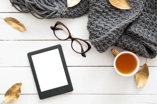 Sonbahar kompozisyonu. Örme eşarp, çay bardağı, gözlük, boş ekranlı tablet ebook ile kadınsı masa masa, ahşap arka plan üzerinde sonbahar yaprakları. Düz yatıyordu, üstten bak. İskandinav, hygge, rahat ev konsepti