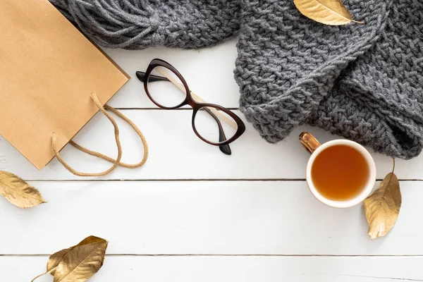 Composizione autunnale. Tavolo da scrivania femminile con sciarpa lavorata a maglia, tazza di tè, bicchieri, shopping bag, foglie autunnali su sfondo bianco in legno. Posa piatta, vista dall'alto. Nordic, hygge, accogliente concetto di casa — Foto Stock