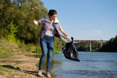 Gönüllü kadın nehrin kıyısında plastik çöp topluyor. Temizlik ortamı kavramı