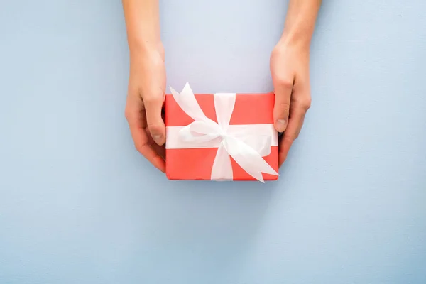 Female's hands holding red gift box with white ribbon bow on pastel blue background. Christmas, New Year, Valentine's day and birthday concept. Minimal flat lay style composition, top view, overhead — Stock Photo, Image