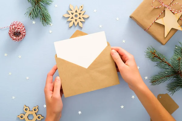 Konfeti, hediye kutusu, köknar ağacı dalları ile süslenmiş pastel mavi arka plan üzerinde kraft kağıt zarf içinde boş boş kart tutan kadın elleri. Noel, Yeni Yıl, kış tatili davetiyesi mockup.