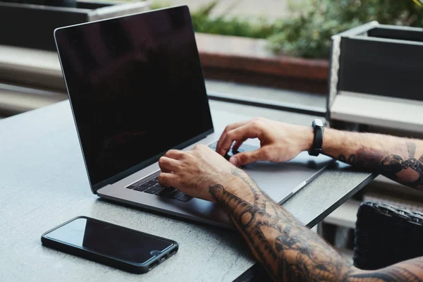 Junger tätowierter Mann, der in einem Café am Laptop arbeitet. Rückansicht von Hipster-Händen mit Tätowierung, die am Coworking-Bürotisch mit Laptop beschäftigt sind. Kerl tippt Text auf Laptop mit leerem Kopierraum-Bildschirm-Attrappe — Stockfoto