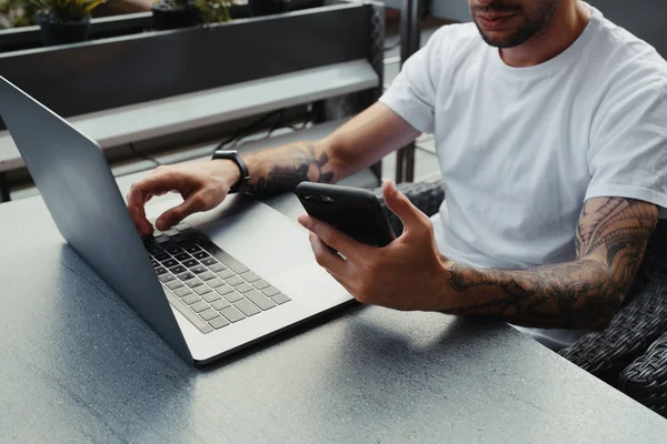 Handsome skäggiga Guy med tatuerade händer klädd vit skjorta med smartphone och bärbar dator i Outdoors kafé, manliga innehav mobiltelefon och skriva text på laptop Keyboard. — Stockfoto