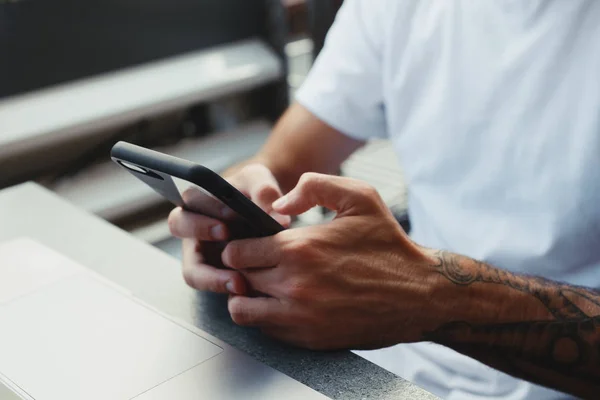 El primer plano de las manos de un hombre es sostener el teléfono inteligente y escribir un mensaje de texto en la pantalla. Hipster chico manos con tatuaje utilizando el teléfono móvil, freelancer leer noticias en Internet, comprobar el correo electrónico, navegar por Internet — Foto de Stock