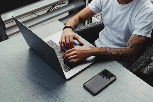 Stilig kille med tatuerade händer klädd vit skjorta arbetar på laptop, manliga skriva text på anteckningsblocket tangentbord. Frilansare arbetar med nya Startup projekt i City café med gratis trådlös zon — Stockfoto