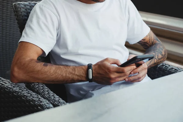 Manos de tatuaje sosteniendo el teléfono móvil. Hombre guapo vestido camiseta blanca sentado en la cafetería y el uso de teléfono inteligente en las manos, navegar por Internet, charlando, escribiendo mensaje. Foto de calle, estilo hipster — Foto de Stock