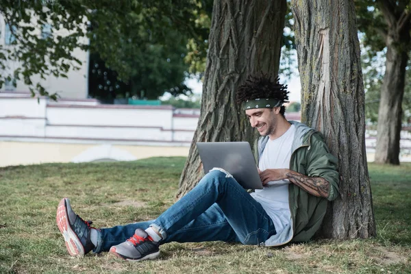 Handsome Young International student sitter på gräs i Public Park, skriva textmeddelanden på tangentbordet i modern bärbar dator, chatta i sociala nätverk. Freelancer med anteckningsbok för distansarbete — Stockfoto