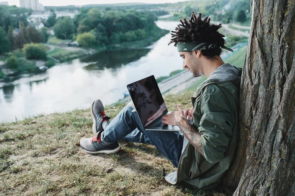 Young tusenåriga student med dreadlocks sitter på gräs och använder bärbar bärbar dator utomhus i offentlig park. Frilansare använda anteckningsbok för distans jobb, distansarbete, e-Learning — Stockfoto