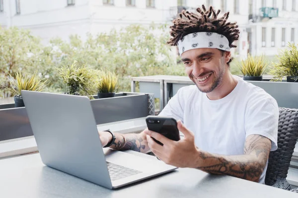 Leende unga manliga frilansare skriva på bärbar dator medan du läser inkomst meddelande på smartphone, positiv hipster kille kontrollera anmälan på mobiltelefon keyboarding på anteckningsbok i Outdoors Café — Stockfoto