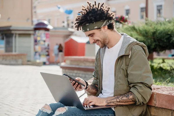 Side View Shot av en stilig man med smartphone medan du arbetar på laptop utomhus, lycklig kille med afrikansk dreadlocks frisyr kontrollera anmälan, läsa meddelande, synkronisera data via WiFi — Stockfoto