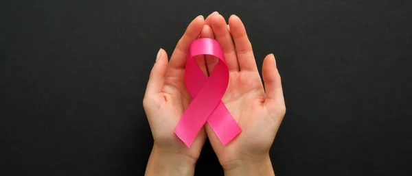 Breast cancer awareness symbol pink ribbon in woman hands on black background. Breast cancer awareness campaign banner.