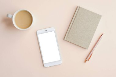 Boş ekranlı cep telefonu modeliyle minimal kadınsı ofis masaüstü, bir fincan kahve, kağıt defter ve bej arka planda kalem. Düz konum, üst görünüm, metin için boşluk.