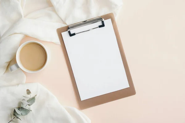 Home office desk with blank clipboard, cup of coffee, blanket and eucalyptus leaf. Modern feminine workspace, minimal style. Flat lay, top view.