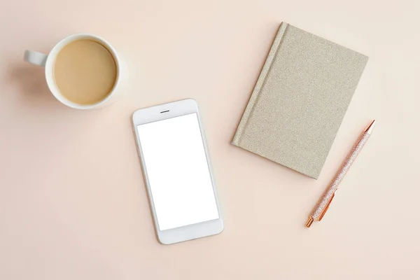Minimal feminine office desktop with mobile phone mockup with blank screen, cup of coffee, paper notebook and pen on beige background. Flat lay, top view, space for text.