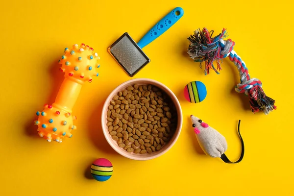 Toys for cat and bowl with dry food on yellow background top view. Pet care and training concept.