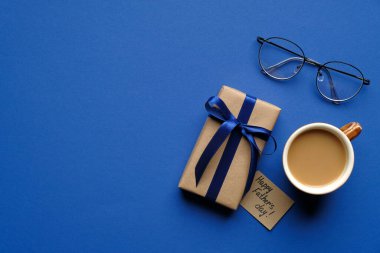Mutlu Babalar Günü konsepti. Üst manzara hediye paketi, mavi kurdele, tebrik kartı, bir fincan kahve ve bardaklar mavi masada. Babalar Günü afiş şablonu.