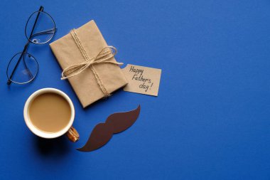 Babalar günün kutlu olsun. Hediye kutusu, tebrik kartı ve bir fincan kahve, bıyık ve mavi arka planda gözlüklü düz kompozisyon. Babalar günü kart şablonu.