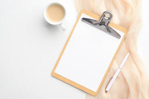 Feminine workspace with clipboard with empty paper note, cup of coffee, beige scarf and pen on white background. Wedding planner, checklist, to do list mockup.