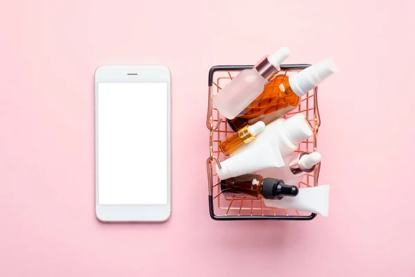 Buy cosmetics online concept. Flat lay mobile phone with blank screen mockup and shopping basket full of cosmetics products on pink background. Flat lay, top view.