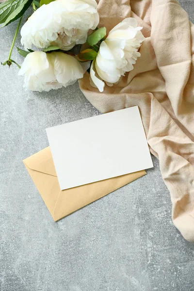 Romantic letter concept. Blank white card with kraft paper envelope, beige cloth and peony flowers on concrete stone table. Flat lay, top view. Wedding invitation mockup, greeting card template.