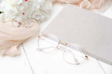 Kağıt defter, bardaklar, şakayık çiçekleri ve beyaz ahşap masada bej kumaşlı düz kompozisyon. Üst Manzara. Zarif kadınsı çalışma alanı, asgari biçim.