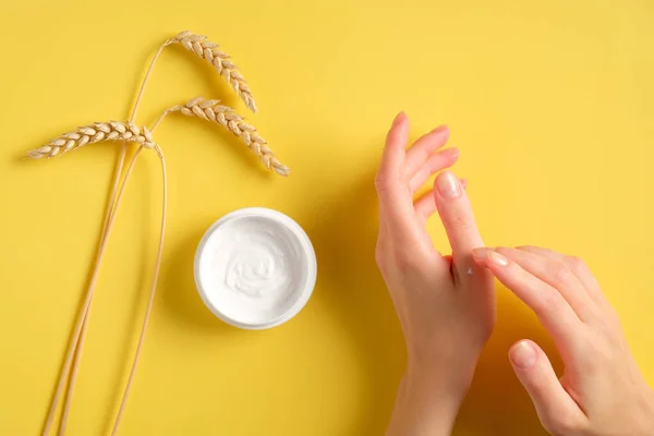 Cosmetic cream on female hands, jars with moisturizer cream and wheat on yellow background. Flat lay, top view. Woman applying organic moisturizing hand cream. Hand skin care concept