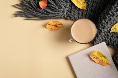Sonbahar düz kompozisyonu, ekose örgü, kahve fincanı, kağıt defter, bej arka planda düşmüş yapraklar. Hygge tarzı kadınsı çalışma alanı, rahat ofis masası. Düz yatış, üst görünüm.
