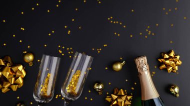 Lüks Noel kompozisyonu. Şampanya şişesi, bardaklar, altın konfeti, zarif Xmas süslemeleri, siyah arka planda konfeti. Düz yatış, üst görünüm.