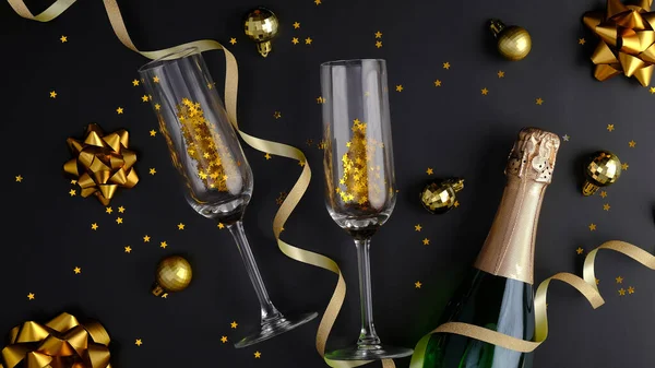 Noel kompozisyonu. Şampanya şişesi, bardaklar, altın toplar, süslemeler ve konfetiyle siyah Xmas geçmişi. Düz yatış, üst görünüm.