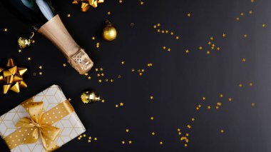 Noel kompozisyonu. Düz yerde şampanya şişesi, hediye kutusu, xmas süslemeleri, süslemeler, siyah arka planda altın konfeti. Noel tebrik kartı, bayram afişi.