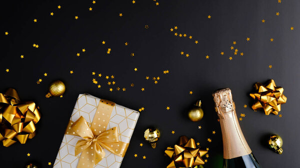 Christmas or New Year Eve celebration concept. Luxury Christmas composition with champagne bottle, gift box, golden decorations, confetti on black background. Flat lay, top view.
