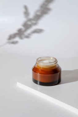 Beyaz kürsüde kehribar cam kavanozda doğal organik nemlendirici krem. Arka planda kurumuş çiçeğin gölgesi.