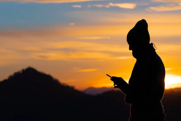 Silhouette of a male hiker with knit cap looking at his mobile phone on a beautiful sunset background. Hiking, modern technology and nature concepts.