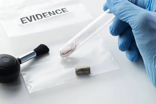 Closeup of forensic scientist's hand in blue rubber glove holding glass tube over evidence bag with 9 mm bullet case next to blower brush. Crime, forensic science, investigation and violence concepts.