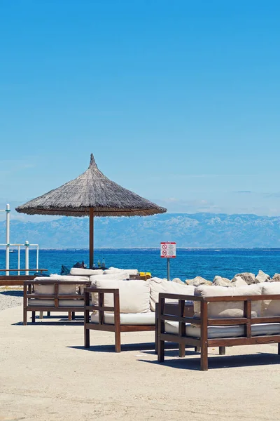 Wooden lounge bar benches with straw umbrella by Adriatic sea in Croatia on a clear sunny day. No dogs and no nudity allowed sign. Vacation, tourism, travel and summer concepts