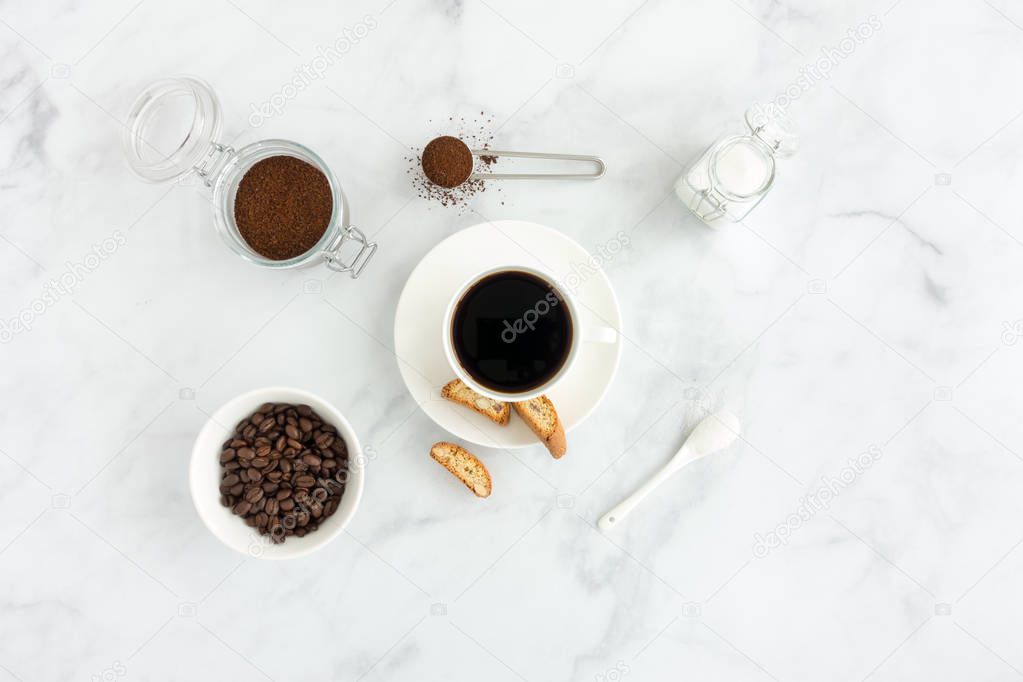 Black coffee in a cup with cantucci, white sugar, ground coffee and coffee beans on white marble background.