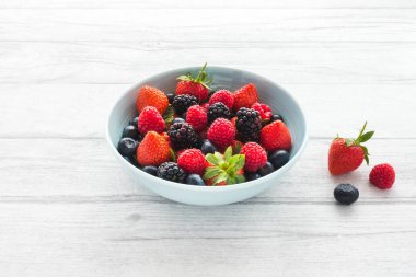 Çilek, ahududu, böğürtlen ve yaban mersini kopya alanı ile beyaz ahşap zemin üzerinde mavi bir kase. Arkadan aydınlatmalı.