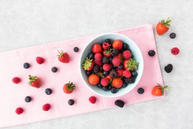 Taze yaz meyveleri pembe kumaş ve kopya alanı ile gri arka plan üzerinde mavi bir kabın içinde çeşididir. Üstten görünüm.