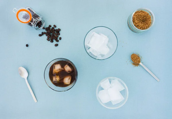 Glasses of cold brew coffee, coffee beans, sugar and a bowl filled with ice cubes on blue background. Top view.