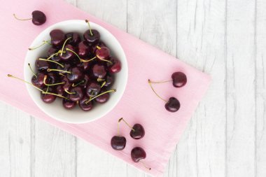 Fresh cherries with stalks in a white porcelain bowl on a pink fabric and white wood. Top view.