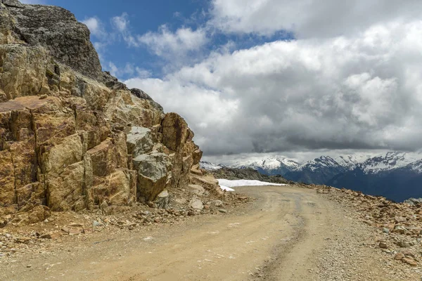 country road, a rocky road in the rocky mountains, near the rock. A blue sky with thick clouds and mountains covered with white snow in the background