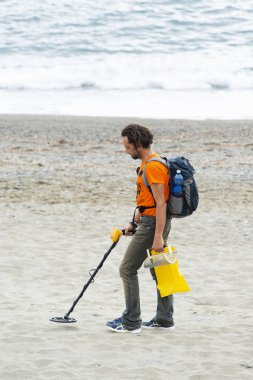 Savona, Italy, October 25, 2018: The man is walking on the beach with a metal detector in one hand and a sieve in the other. He is trying to find jewels and coins, he has a backpack and a yellow bag clipart