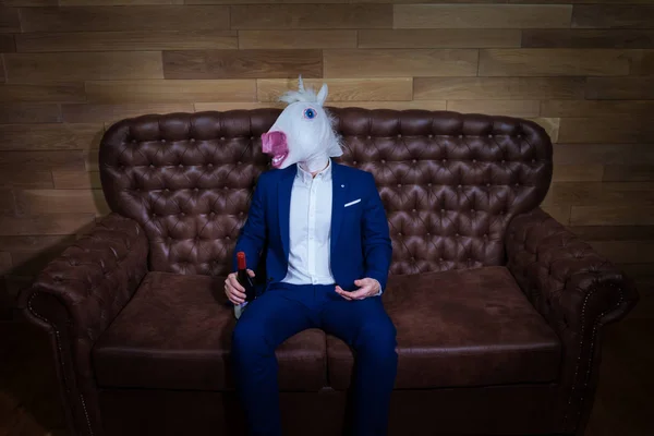 Unicornio divertido en traje elegante se sienta en el sofá con botella de vino —  Fotos de Stock