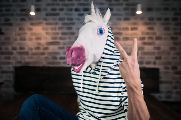 Freaky ung man i komisk mask sitter på sängen i eleganta lägenheter. Rolig unicorn — Stockfoto