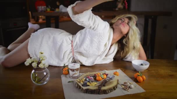 可爱的金发女郎穿着浴袍躺在桌子上 靠近木托盘 在时尚的厨房里摆着煎饼和水果 年轻的昏昏欲睡的女人吃浆果 在家享用早餐 — 图库视频影像